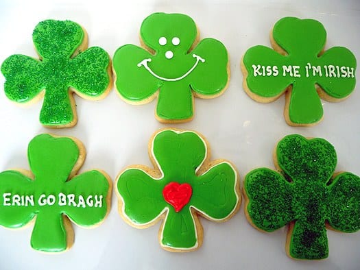 6 shamrock sugar cookies with green icing and piping decorations on a white plate.