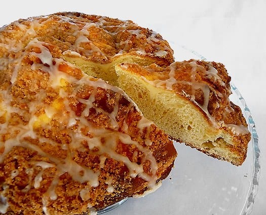 Coffee cake with sweet cheese filling and vanilla icing on a glass cake stand with a slice cut out.