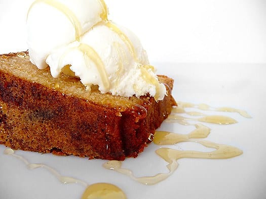 Side view of a slice of honey vanilla sour cream pound cake topped with a scoop of vanilla ice cream and a drizzle of honey on white plate.