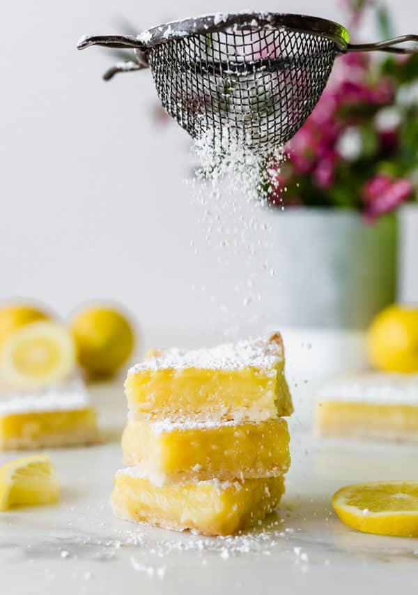A stack of three lemon bars being sprinkled with powdered sugar.