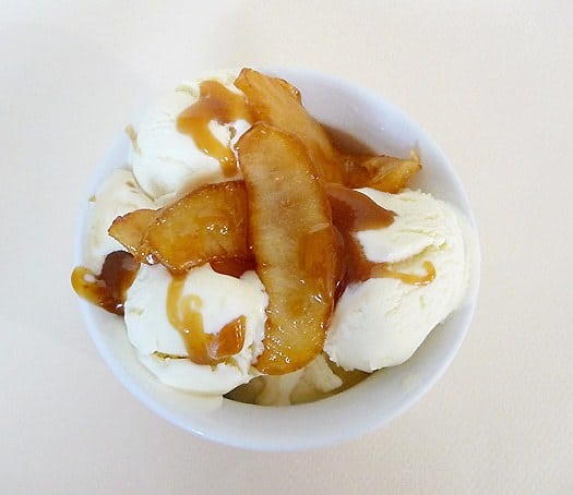 Overhead image of scoops of cinnamon ice cream in a white bowl topped with sautéed apples and caramel sauce.
