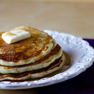Stack of 3 pancakes topped with a pat of butter and maple syrup on a white plate.