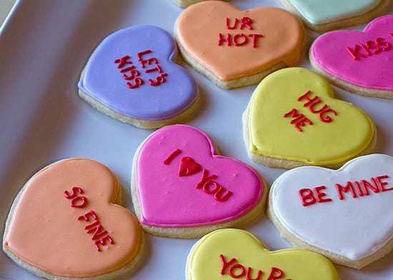 Heart shaped sugar cookies decorated like conversation hearts on a white plate.