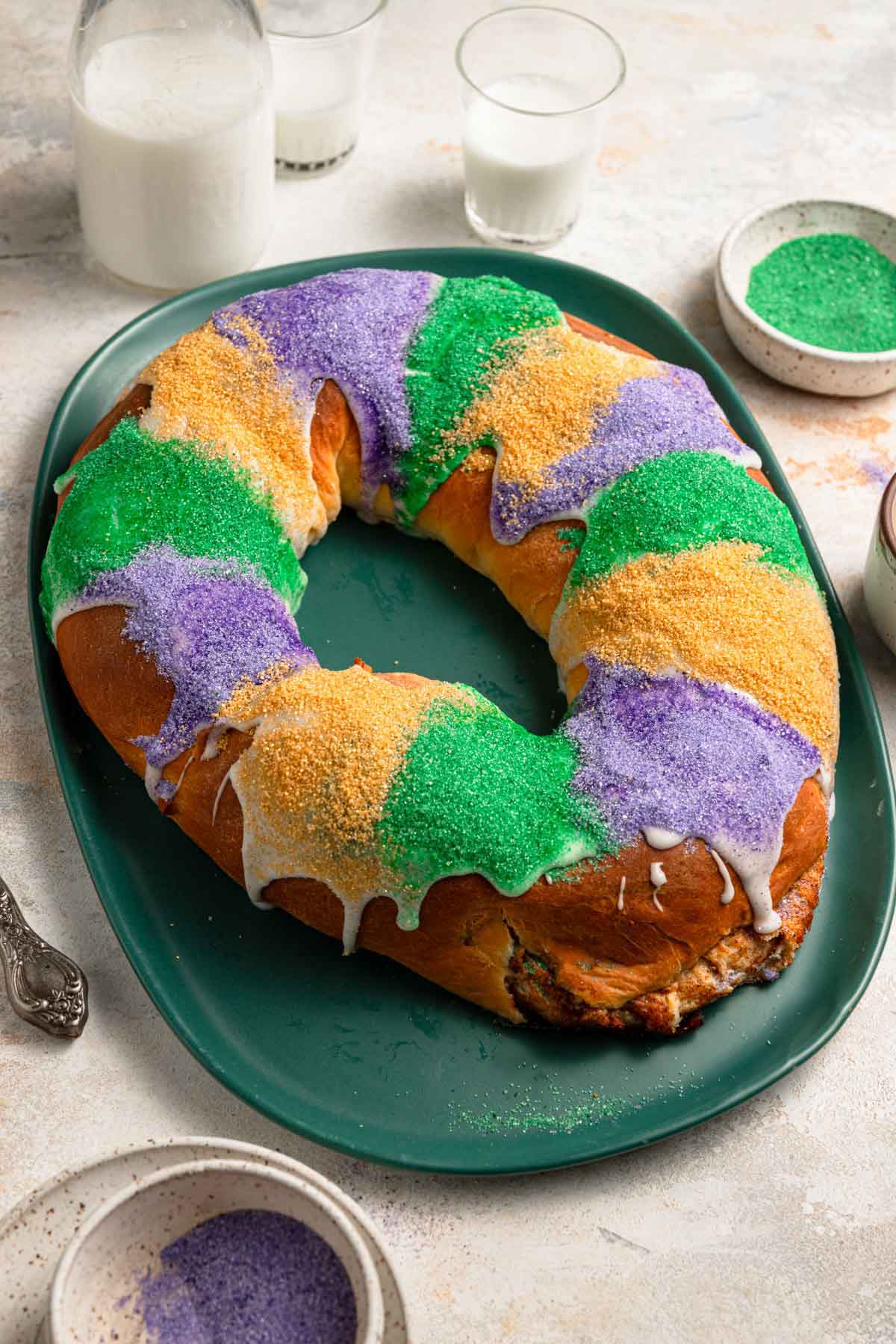 Photo of baked king cake on a green platter iced and decorated with sanding sugar in alternating strips of purple, green, and yellow.