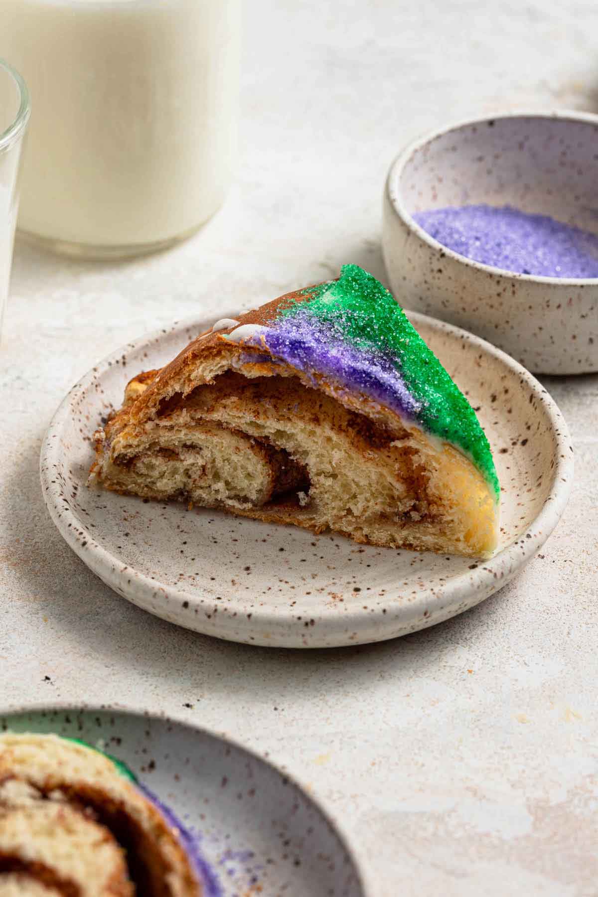 A slice of king cake on a speckled plate with a cup of milk and bowls of colored sanding sugar in the background.