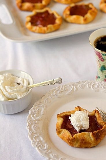 Rhubarb pie tartlet on a white plate with a fork and whipped cream in a white bowl with a spoon.