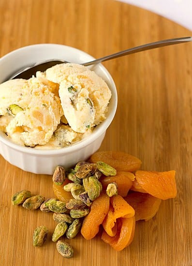 Scoops of dried apricot pistachio ice cream in a white bowl with a spoon with pistachios and dried apricots next to the bowl.
