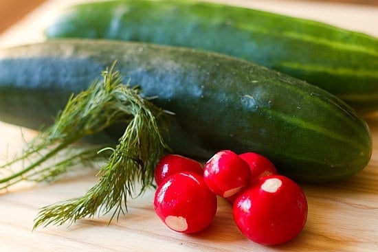 Ingredients for creamy cucumber salad including cucumbers, radishes, and dill on a wood cutting board.