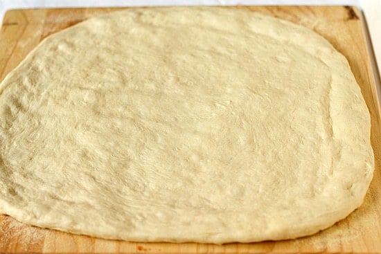 Pizza dough rolled out on a wood cutting board.