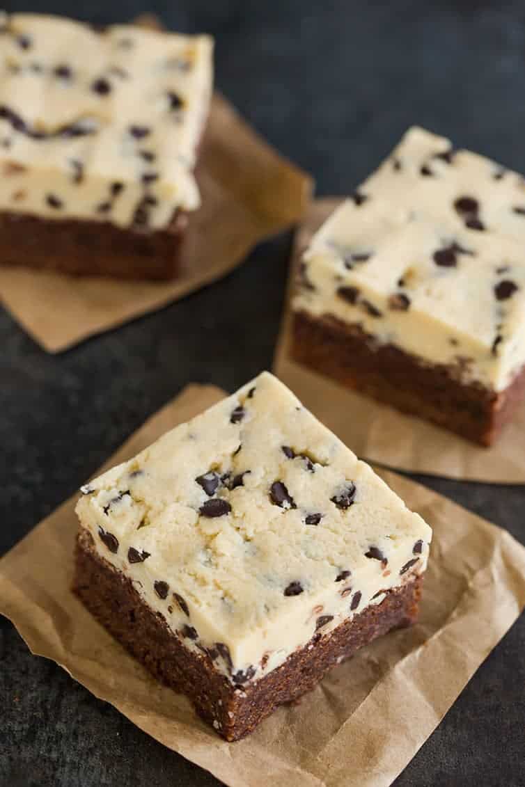 Three chocolate chip cookie dough brownies sitting on squares of parchment paper.