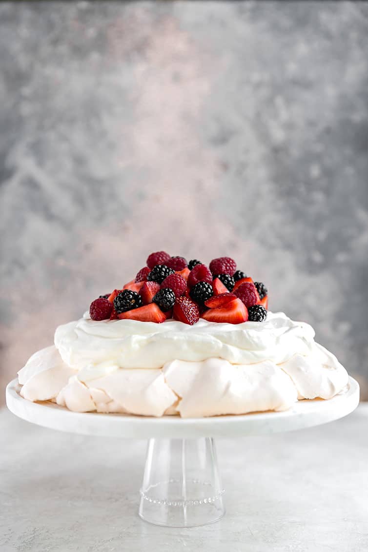 Pavlova on a glass cake plate, with whipped cream and berries.