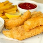 Beer battered cod and french fries on a white plate with lemon wedges, tartar sauce in a white bowl, and ketchup in a white bowl.