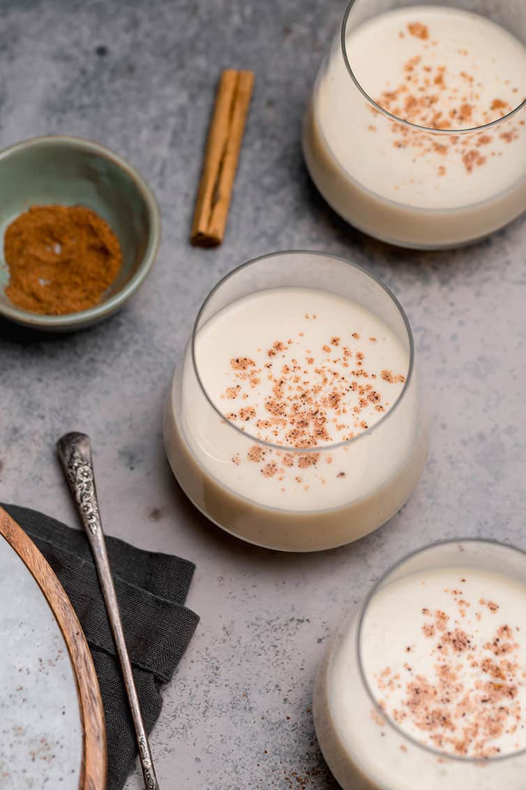 Three glasses of eggnog with a cinnamon stick and a bowl of grated nutmeg.