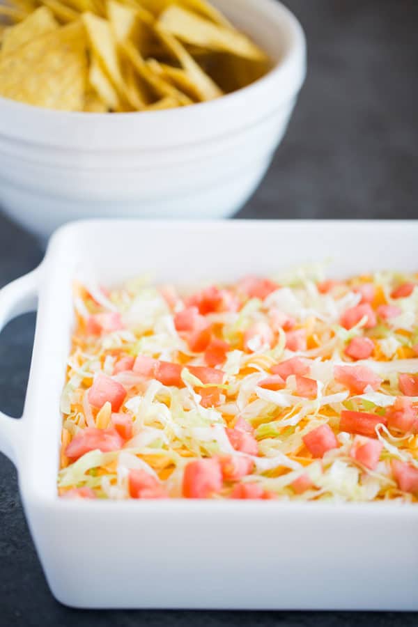 Taco dip in a white casserole dish with a bowl of chips in the background.