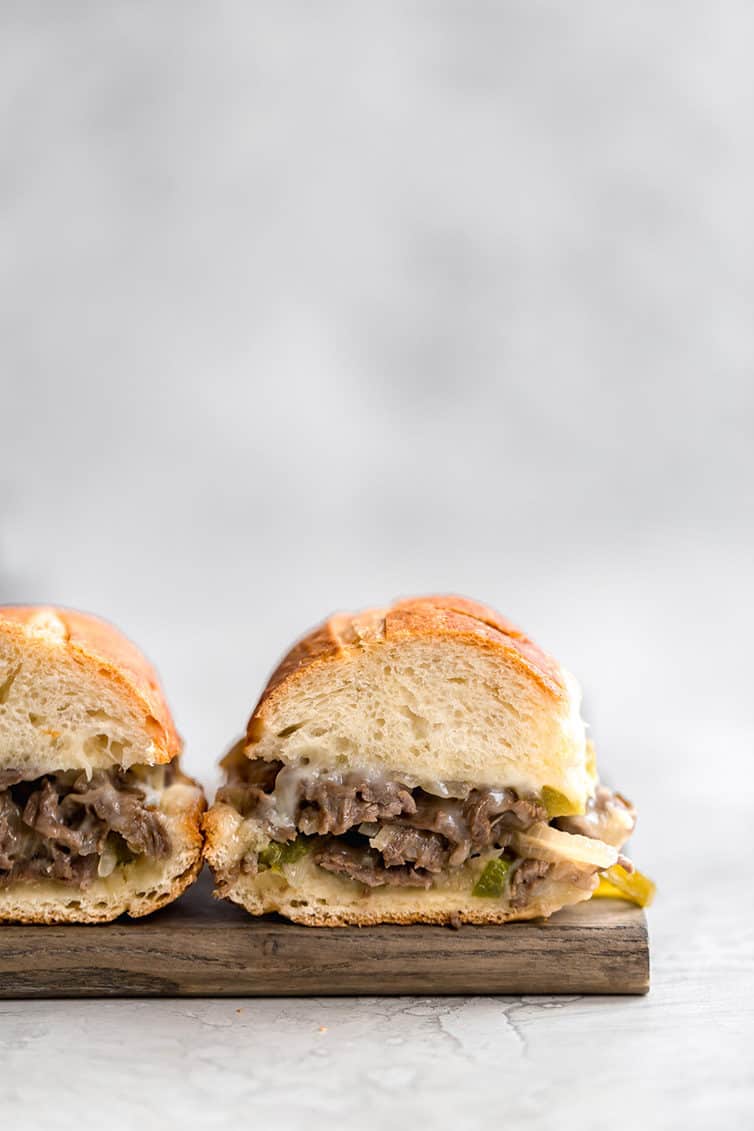 Two halves of Philly cheese steak sandwiches on a wooden cutting board.