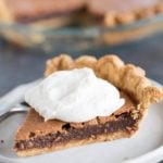 A slice of chocolate chess pie with whipped cream on top in front of the pie plate with the rest of the pie.