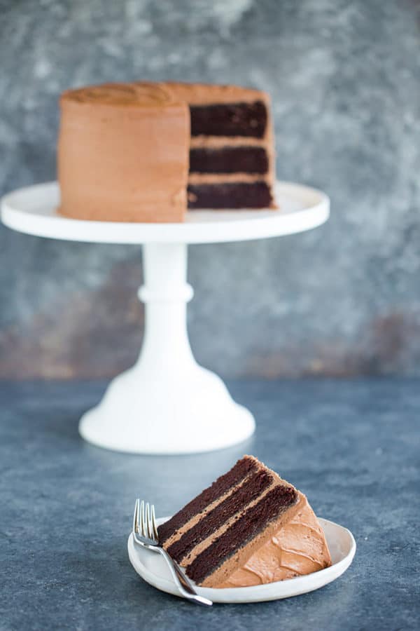 A slice of chocolate cake on a plate in front of the whole cake on a serving platter.