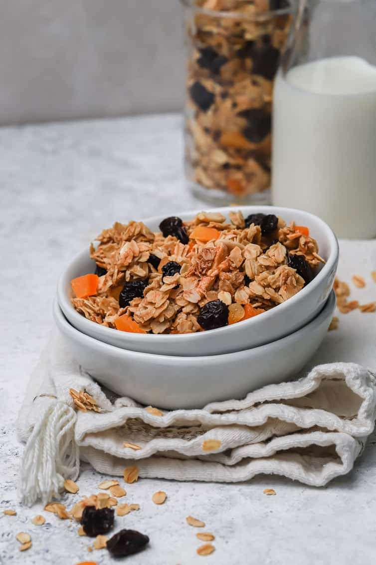 A bowl of granola with a glass of milk and jar of granola in the background.
