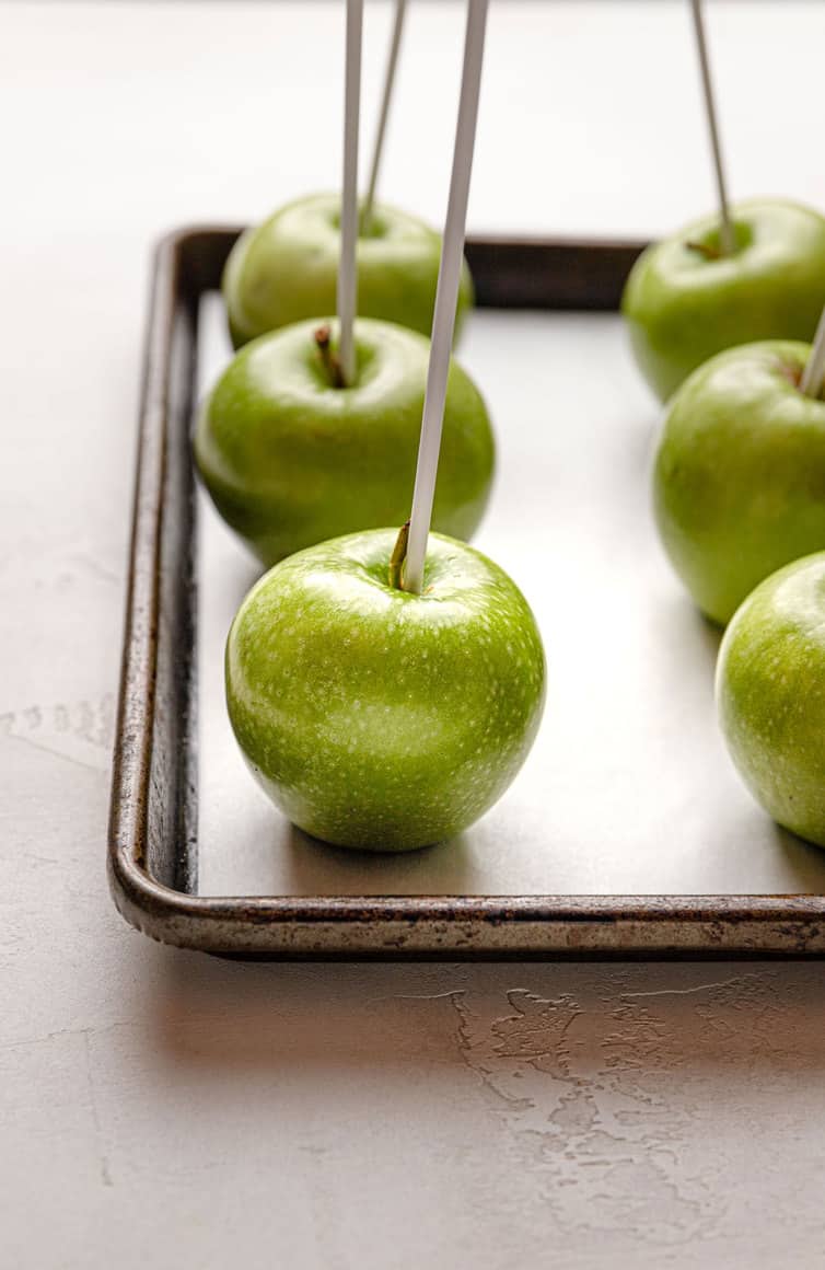 Granny Smith apples with lollipop sitcks inserted in center on a baking sheet.
