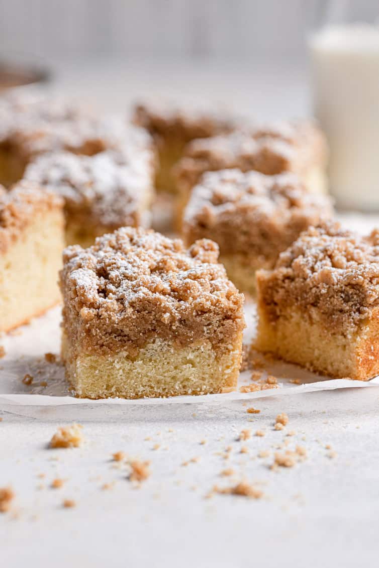 Squares of crumb cake on a surface with glass of milk in background.