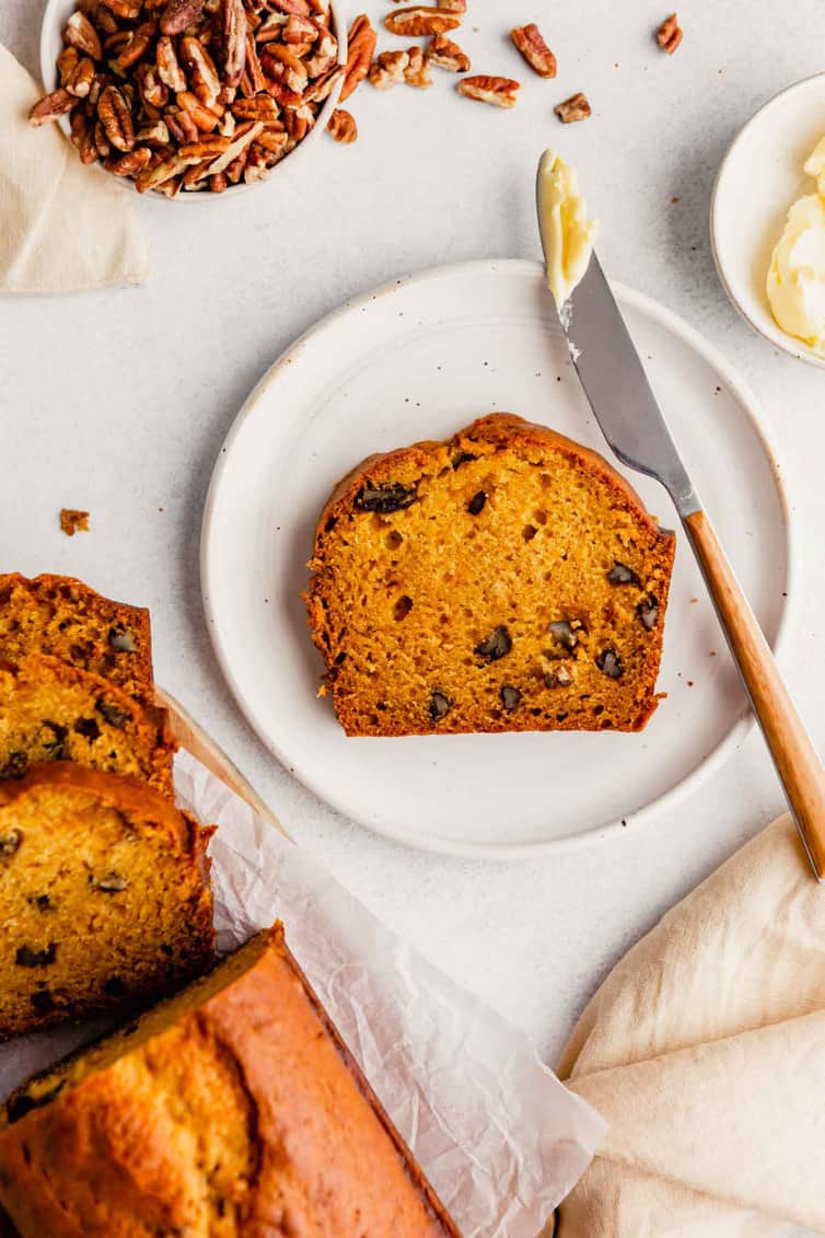 One slice of pumpkin bread on a white plate with a butter knife with butter next to it.