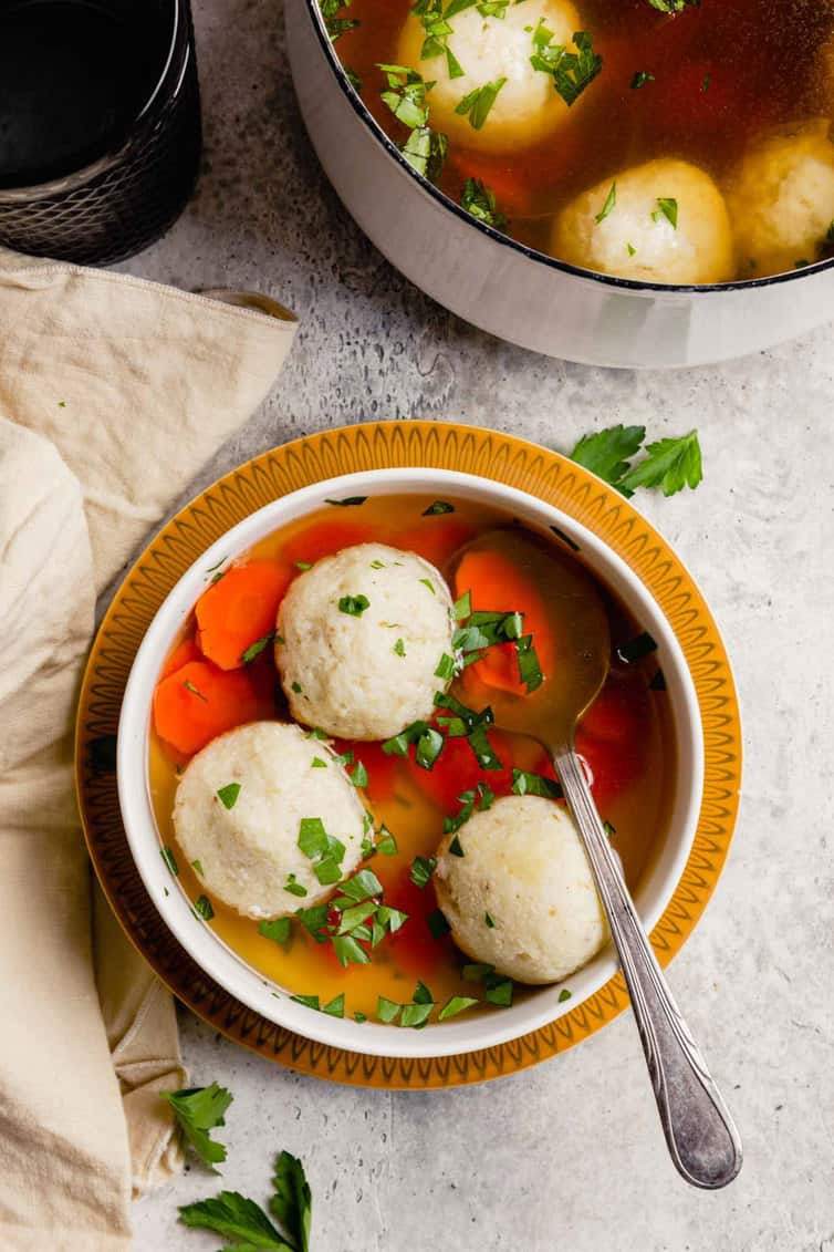 A bowl of matzo ball soup with a spoon and a pot of matzo ball soup in the back.