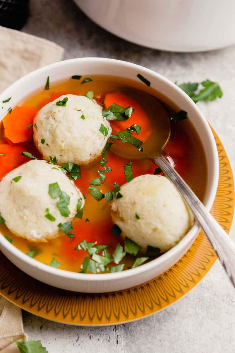 A warm bowl of matzo ball soup on a yellow plate with a silver spoon in the bowl topped with parsley.