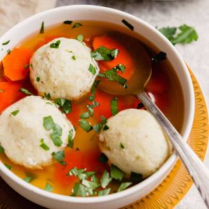 Homemade matzo ball soup in a white bowl with three cooked matzo balls on top with a silver spoon in the bowl.
