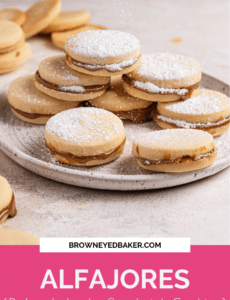 A small white plate with a stack of alfajores dusted with powdered sugar and a pink rectangle at the bottom with the word Alfajores (Dulce de Leche Sandwich Cookies) at the bottom in white.