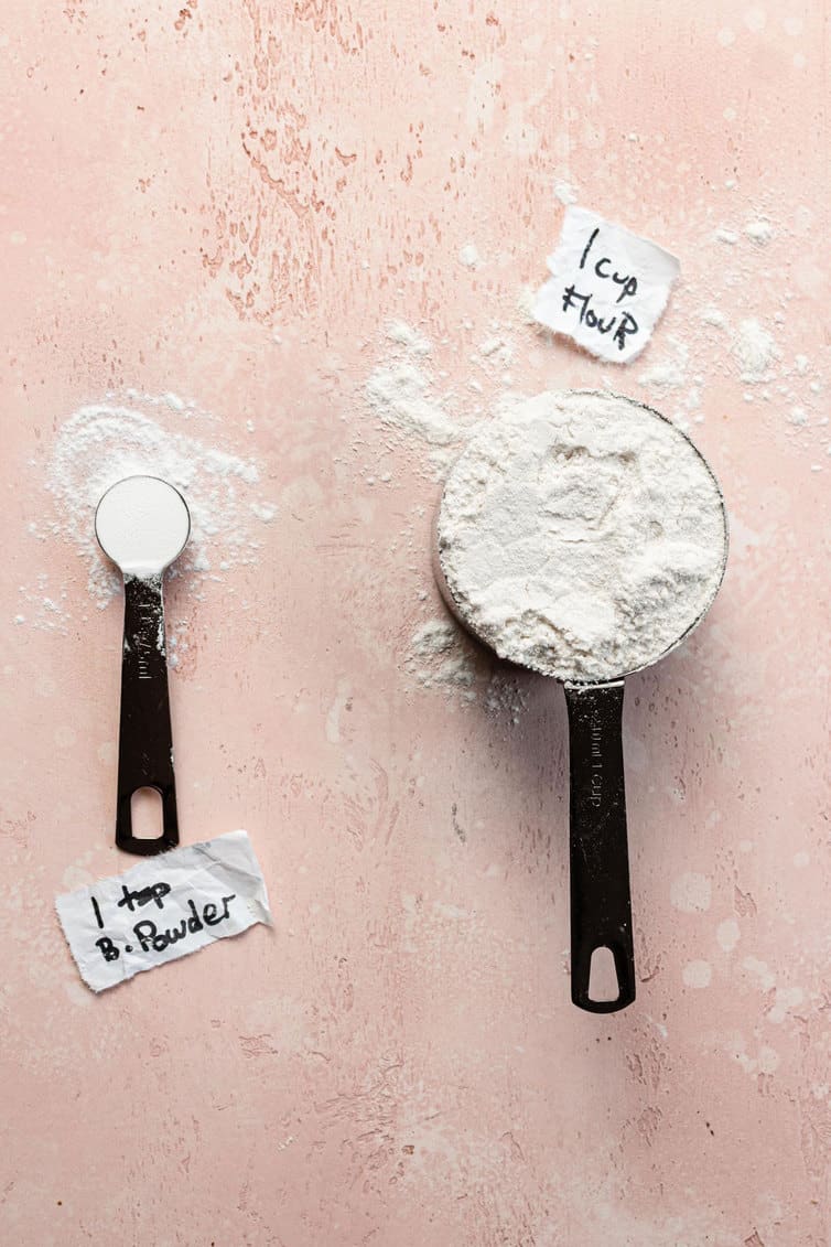 A measuring spoon of baking powder next to a measuring cup of flour.