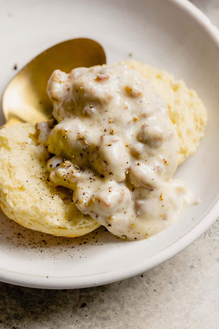A close up shot of sausage gravy on top of an open biscuit on a white plate.