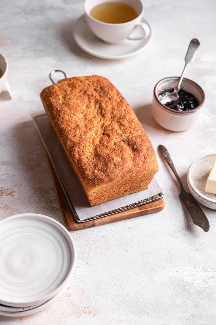 A loaf of english muffin bread on a cutting board with plates to the left and jam in a bowl on the right.