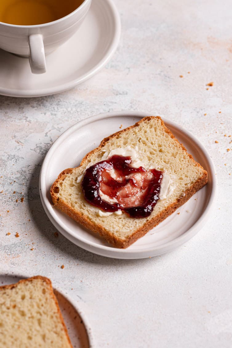 A slice of English muffin bread with butter and jam in front of a mug of tea.
