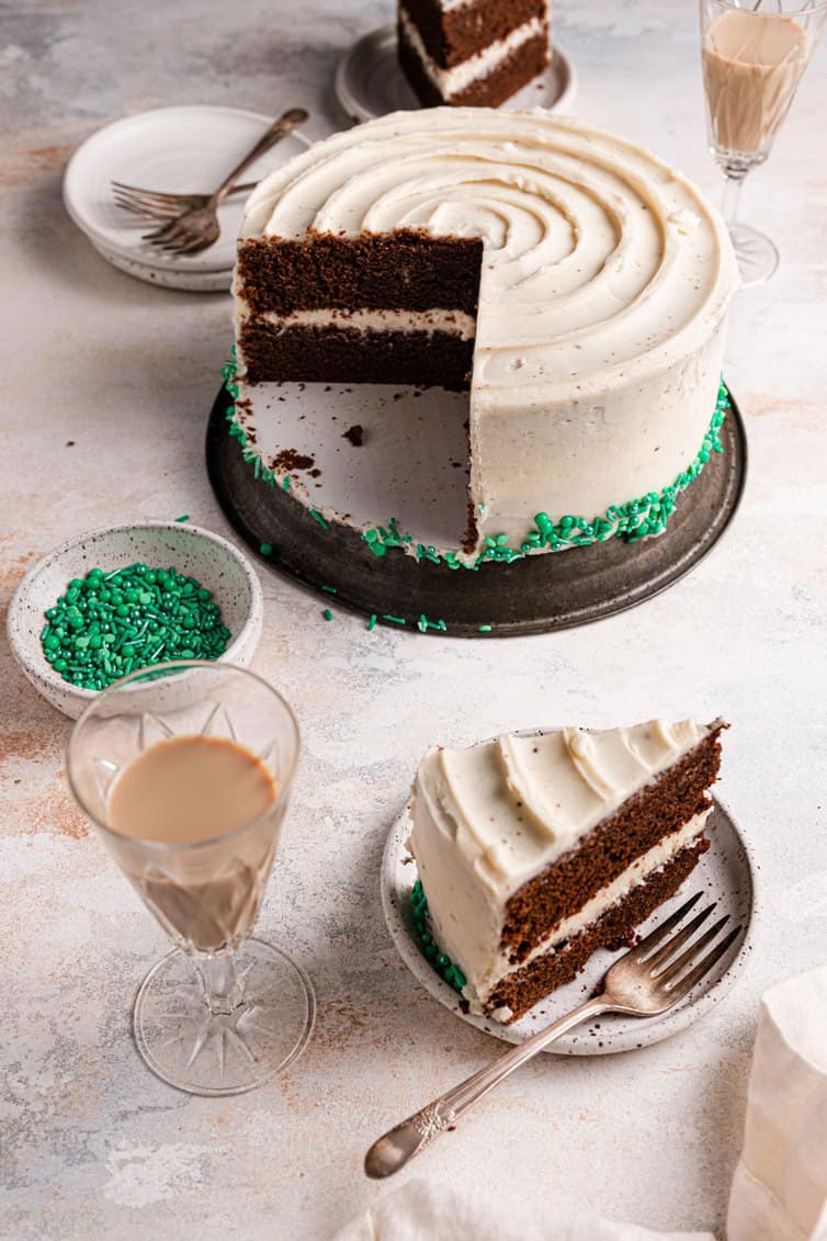 A slice of guinness chocolate cake on a plate with a fork in front of a full cake on a silver stand.