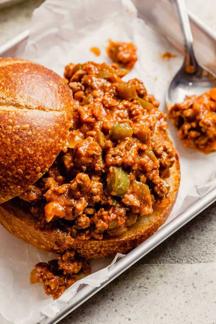 Homemade sloppy joes with the bun top pulled back and a spoon of sloppy joe meat to the right.