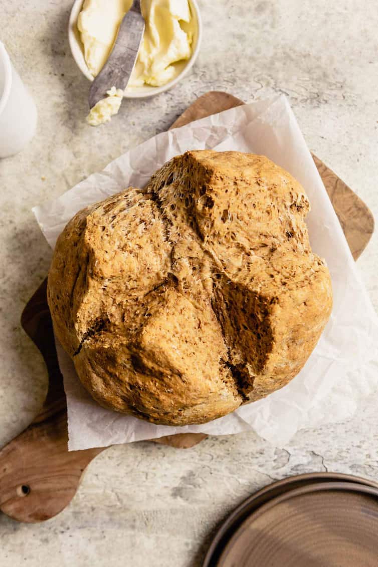 A wooden cutting board topped with parchment paper and a loaf of Irish brown bread with a bowl of butter and a butter knife at the top.