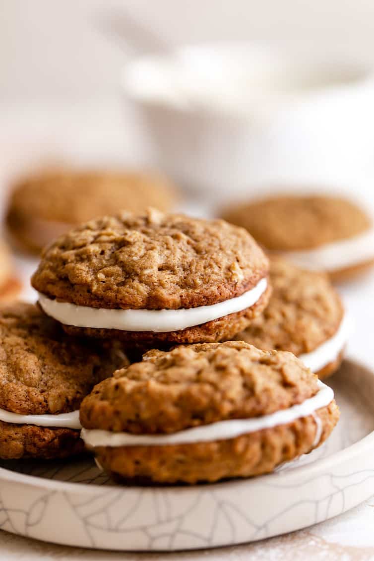 A stack of oatmeal cream pies on a white plate with a white bowl in the back right.