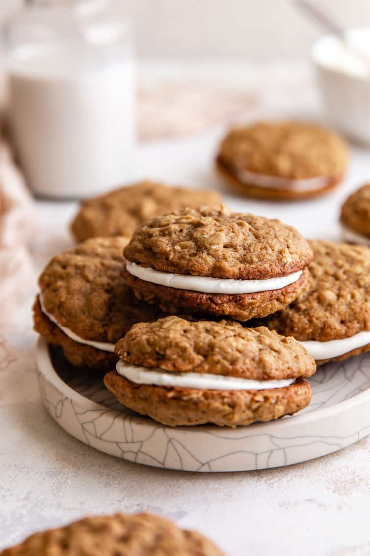 A stack of four oatmeal cream pies on a white plate with a glass of milk in the back left.