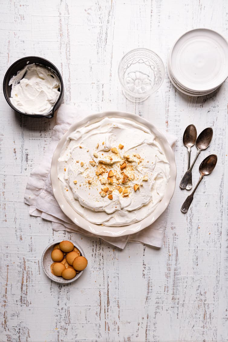 A banana pudding in a pie pan on a towel with spoons on the right a bowl of whipped cream in the top left and a bowl of eggs in the bottom left.