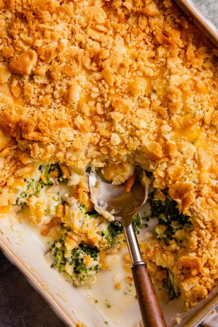 A casserole dish with broccoli casserole with a spoon with a wooden handle and a scoop of broccoli casserole taken out.
