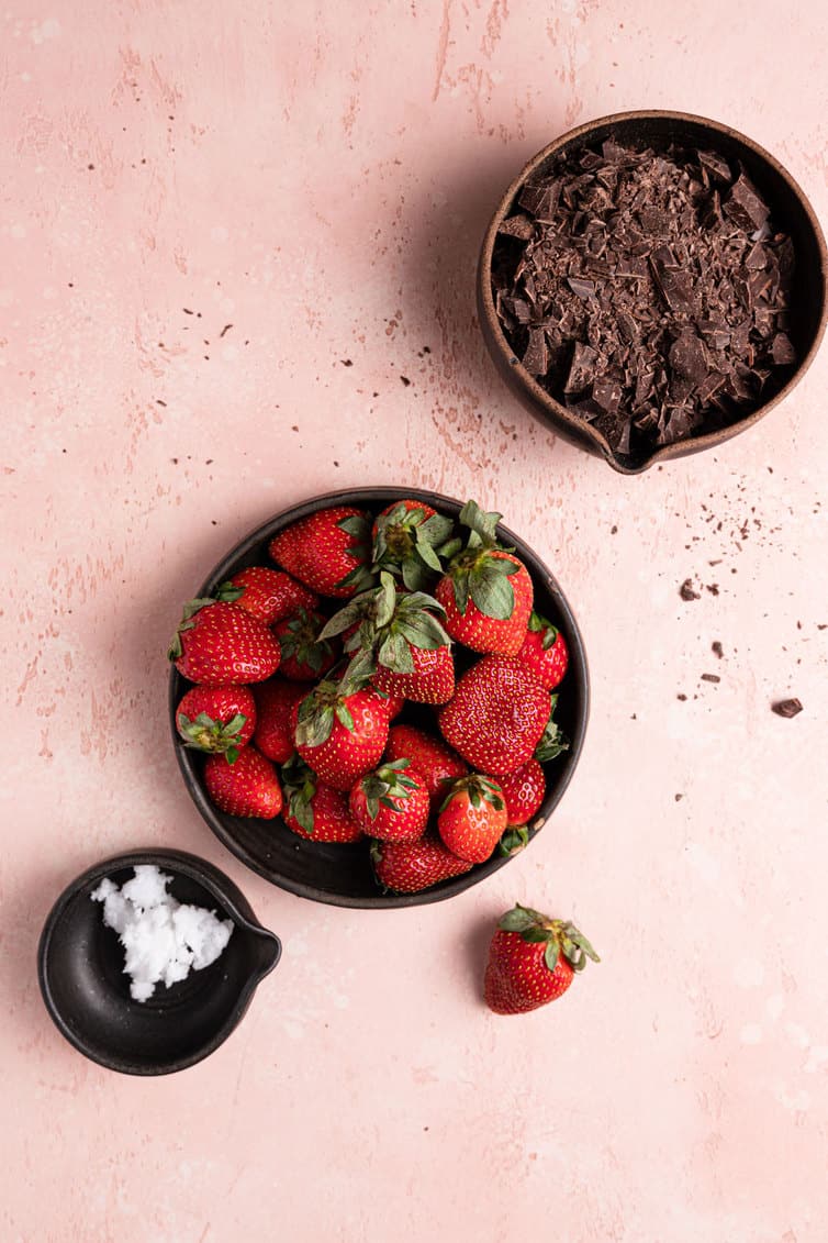 A bowl of chocolate in the top right, a bowl of strawberries in the middle with a strawberry to the side, and a small bowl of coconut oil in the bottom left on a pink counter.
