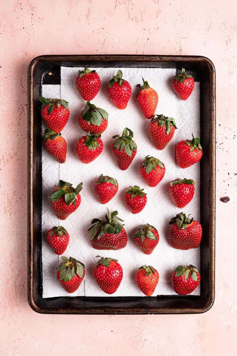 A paper towel lined baking sheet with fresh strawberries on a pink counter.