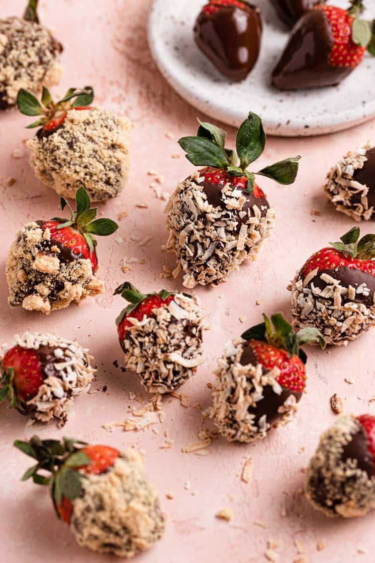 Chocolate strawberries covered in nuts and graham crackers on a pink counter with a small plate in the back with plain chocolate strawberries.