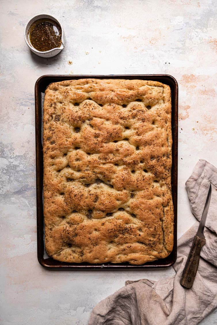 A baking sheet with fresh baked focaccia and a small bowl of herb oil at the top with a bread knife on the right.