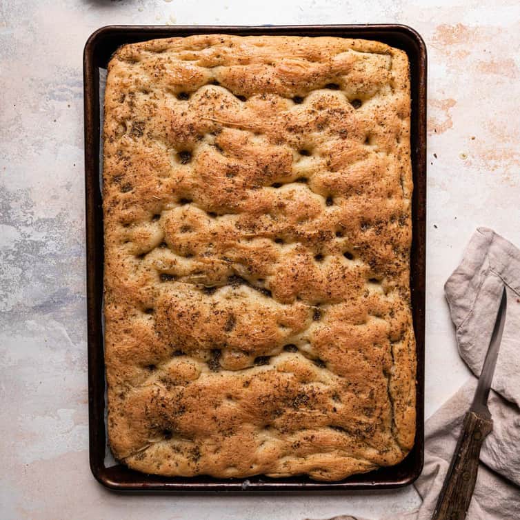 A top down photo of a baking sheet lined with parchment paper and fresh baked focaccia next to a bread knife on the right.