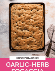A sheet pan with fresh baked focaccia on a white counter with a pink rectangle at the bottom and the words Garlic- Herb Focaccia at the bottom.