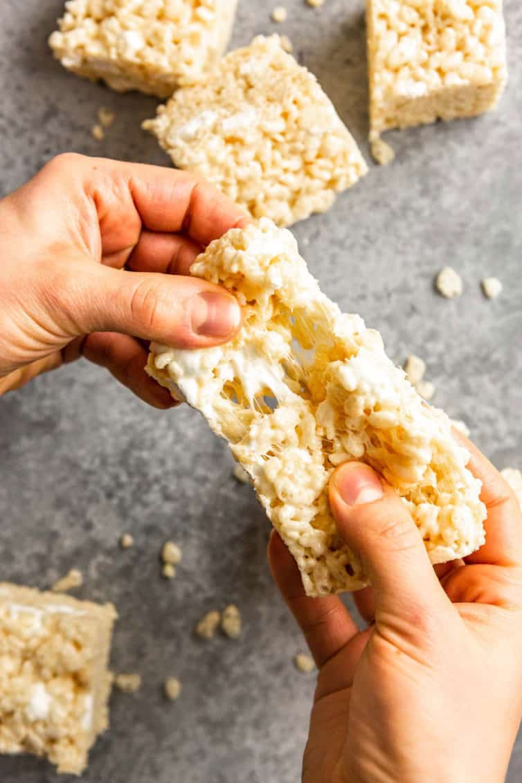 Two hands pulling a rice krispie treat over a grey counter with other rice krispie treats below.