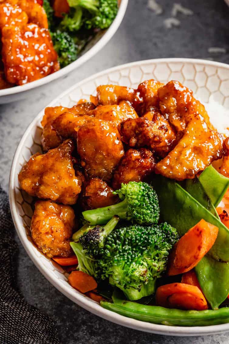 A white bowl with sweet and sour chicken and stir fry vegetables in front of another bowl of sweet and sour chicken on a grey counter.