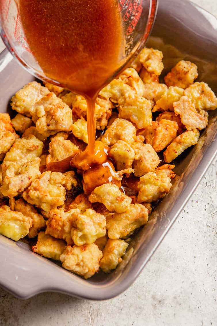 A baking dish filled with fried chicken pieces with sweet and sour sauce being poured over.