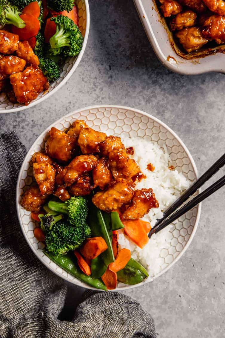 a fresh bowl of sweet and sour chicken with chopsticks on a grey counter with another bowl of sweet and sour chicken in the top left and a pan of the chicken in the top right.